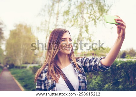 Beautiful blonde Caucasian teenage girl taking a selfie with smartphone in park in spring. Cute happy young woman photographing herself. Horizontal, retouched, filter, copy space.