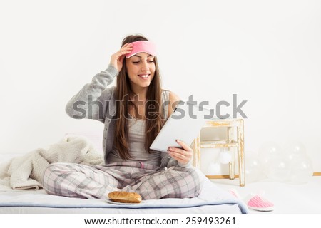 Young woman in bed in morning with digital tablet. Happy teenage girl in pajamas sitting in her bed at home with tablet, croissant for breakfast. No retouch, natural light, horizontal.