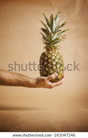 Male hand holding pineapple against brown background. Closeup of hairy man\'s hand holding fresh and ripe pineapple against brown background. Vertical, retouched, warm colors.