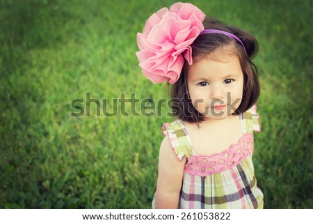 Adorable toddle girl with big pink bow headband and cute romantic pink and green dress smiling looking at camera against green grass background. Horizontal, retouched, filter, copy space.