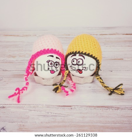 Closeup of two funny Easter eggs with colorful knitted yellow and pink handmade beanie hats and hand drawn cute faces. Square format, matte filter.