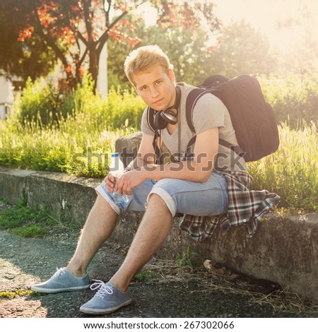 Young hipster man with backpack and headphones in park sitting relaxing holding a bottle of water. Square format, retouched, color filter applied.