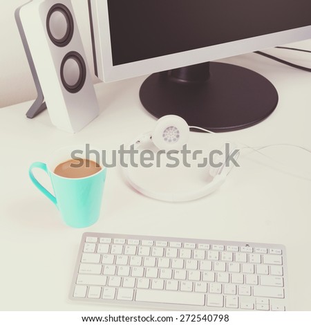 Modern white minimalist home office. Closeup of modern wireless keyboard, turquoise coffee cup, headphones, monitor and speakers. Square format, matte filter.