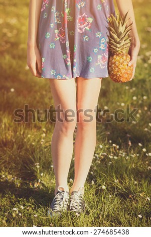Closeup of young unrecognizable slim woman\'s slim legs. Woman in blue dress and canvas shoes holding pineapple standing on field of daisies. Vertical, retouched, matte filter applied.