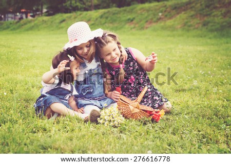 Three cute little girls on picnic in park on sunny summer day. Three little sisters hugging playing outdoors in nature. Horizontal, retouched, filter.