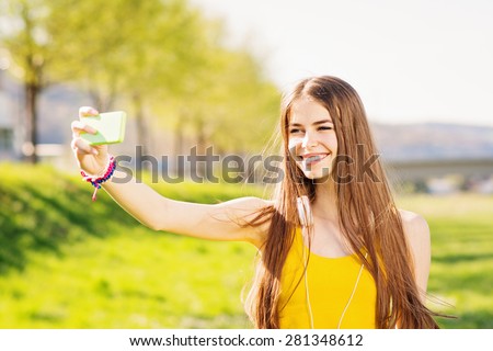 Closeup of beautiful teenage girl in yellow tank top taking a selfie on smart phone outdoors in summer. Young woman photographing herself smiling. Mild retouch, vibrant, natural light, horizontal.