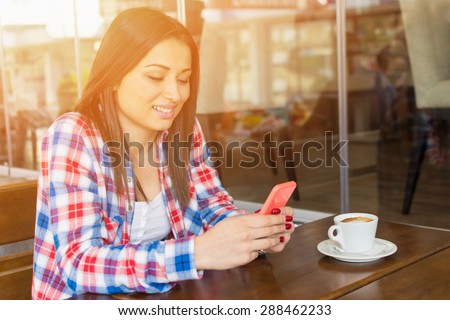 Beautiful young mixed race woman in red and blue plaid shirt with smartphone and espresso coffee texting sitting in cafe bar. Cute teenage girl in coffee shop. Horizontal ,retouched.