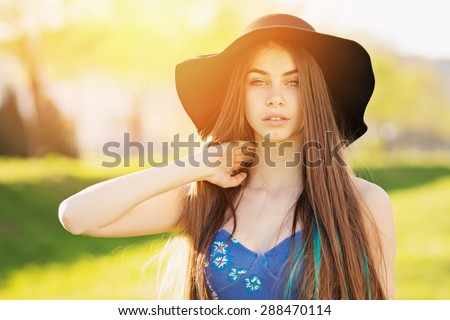 Closeup of gorgeous young woman in blue dress and fedora hat. Beautiful girl posing outdoors on sunny summer day. Horizontal, retouched, vibrant colors.