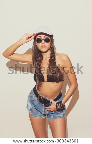 Modern young woman in crochet bikini top, denim shorts, white cap and sunglasses. Modern brunette in summer outfit with braids. Matte filter, studio shot, intentional shadow on the wall.