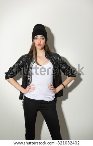Beautiful urban trendy teenage girl in white shirt, black leather jacket, black beanie hat and jeans pouting posing looking at camera with hands on hips. White background, vertical, mild retouch.