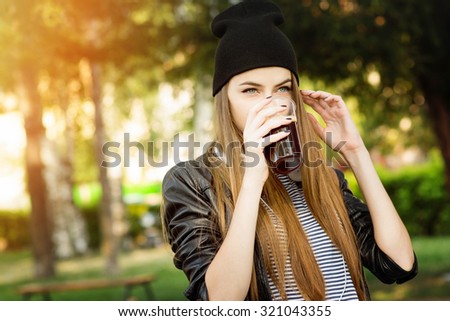 Closeup of beautiful urban cool teenage girl in black leather jacket and black beanie hat drinking dark beer or coffee outdoors in park. Retouched, horizontal, vibrant colors.