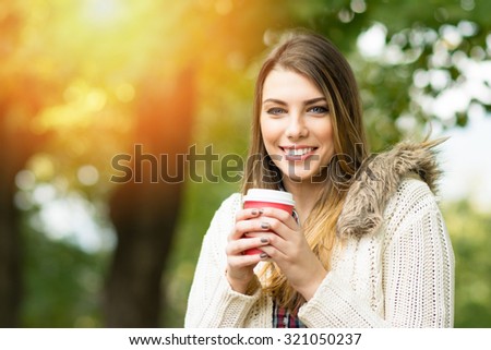 Beautiful young woman in park in autumn smiling holding cup of takeaway coffee. Happy blonde teenage girl outdoors in fall wearing beige sweater. Retouched, horizontal, vibrant colors.
