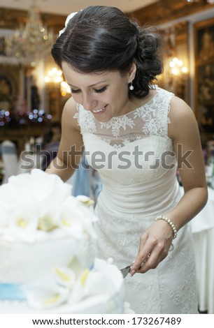 Smiling beautiful bride in an elegant wedding dress bending forwards cutting the wedding cake at the reception