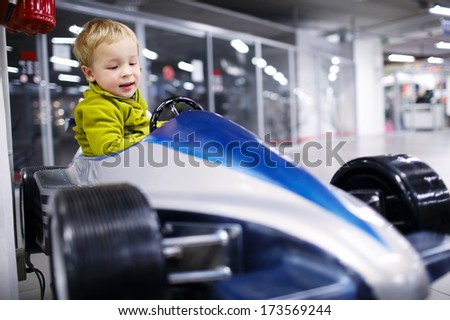Little boy enjoying himself driving an elevated model racing car in an indoor mall or car showroom.