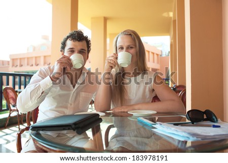 Young couple drinking coffee on an outdoor balcony sitting together with their cups raised to their mouths as they relax and enjoy the view and the summer sunshine