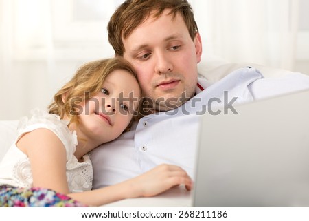Father and little daughter on the sofa at home. Father holding laptop,  girl using it leaning on dads shoulder