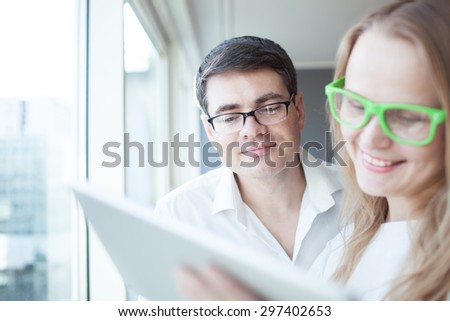 Happy young businessman and businesswoman in glasses using touch pad by the window in light office