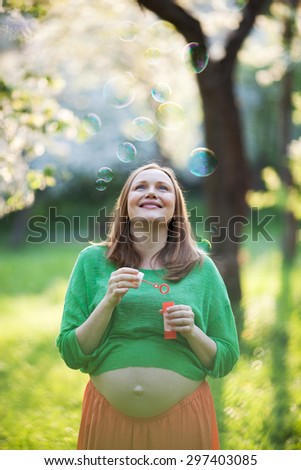 Cheerful young pregnant woman enjoying worry-free time in the park. She looking at flying bubbles on background of blurred nature scene on sunny day