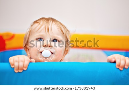11 months Baby boy with dummy in mouth reaching out of his bed holding the edge with hands and looking up.