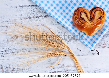 Rustic home-made heart shape freshly backed cinnamon roll on blue checkered cloth over white painted board or table. Above view