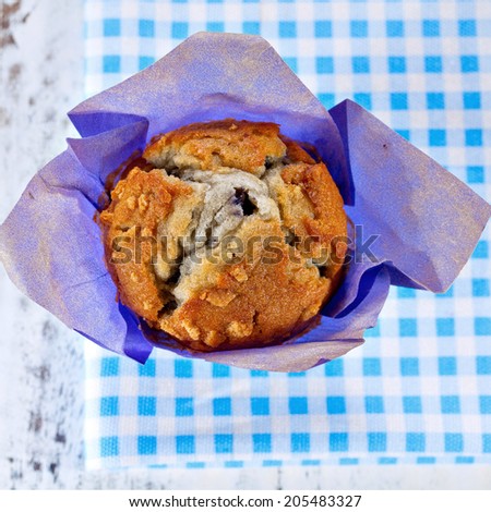 Homemade blueberry muffin wrapped in paper on blue checkered cloth. Top point of view