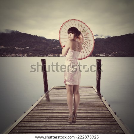 Young pretty chinese girl dressed in traditional pink national dress posing on old wooden pier with  Pella town and misty Italian Alps mountains on background. Isola San Giulio on Lago d\'Orta