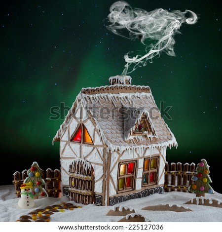 Decorative gingerbread house with lights inside and smoke coming out the chimney on Northern lights background with moon and stars . Rural Christmas night scene