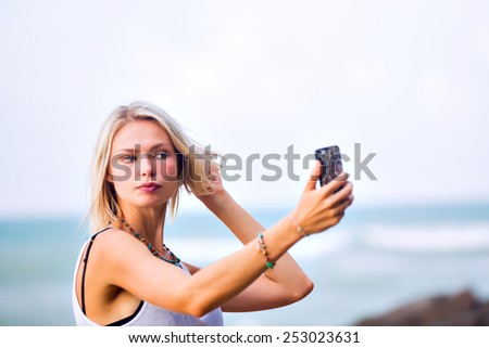 Beautiful young blonde woman taking a selfie on smart-phone outdoor at the rocky sea shore. Trendy fashion female model dressed in white top