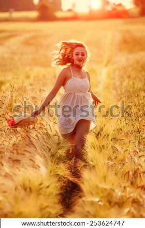 Happy young blonde Scottish girl in white dress with straw hat running through the golden wheat field on sunset light behind her
