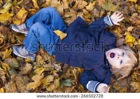 Small blond boy with blue eyes lays on bed of autumn fallen leaves with open mouth