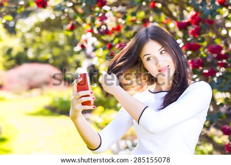 Beautiful young woman with long straight dark hair posing to herself for a selfie with her smart phone in the garden