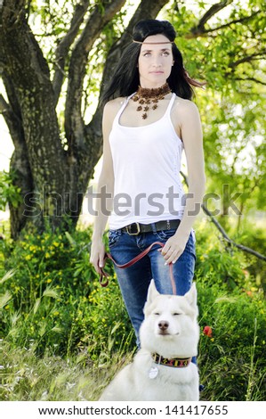 Beautiful injun woman and white husky outdoors.Bellicose brunette woman keeping siberian husky dog on leash, standing in green forest in shadow near the tree.Hippie on a walk with dog