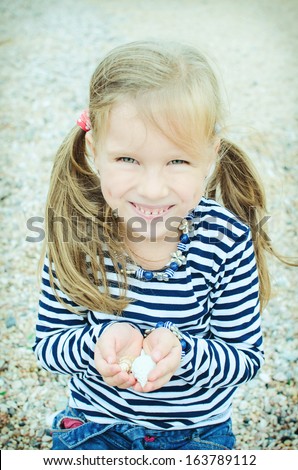 adorable girl on beach in cold season. funny little girl in vest holding seashells in palms. Smiling kid collecting seashells at beach