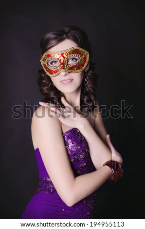 Beautiful fashionable woman in exquisite mask in studio. Fashion portrait of young sexy alluring masked woman girl in elegant evening lilac dress posing on black background