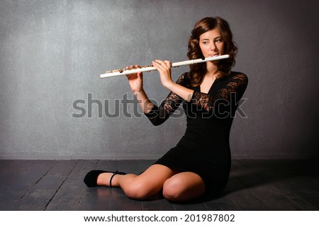 Beautiful young woman playing flute sitting on the floor in studio. Alluring flutist in black evening dress with hairstyle makeup posing with flute on gray background