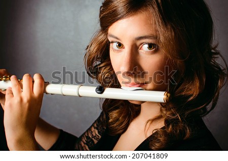 Beautiful young woman playing flute in studio. Alluring flutist in black evening dress with hairstyle makeup posing with flute on gray background