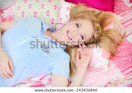 Beautiful fashion woman in trendy casual clothes lying on pink bed linen indoors. Blond teenager girl beauty model in blue shirt with hairstyle and natural makeup. Alluring happy girl.