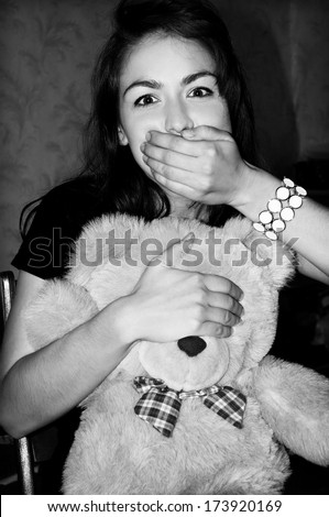Black and white  photo. Portrait of beautiful girl with dark hair. Smile, a kind face. Teddy bear, children\'s toy, bow in the box. White bracelet on his arm, costume jewelery