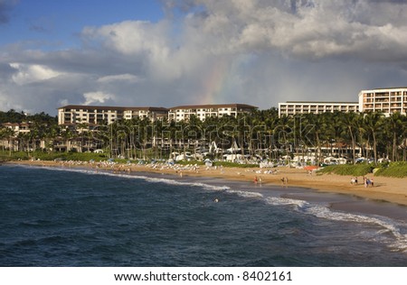A small rainbow crowns hotels along a palm tree lined golden beach and blue ocean.