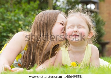Two sisters lying on the grass, one kissing the other on her cheek.