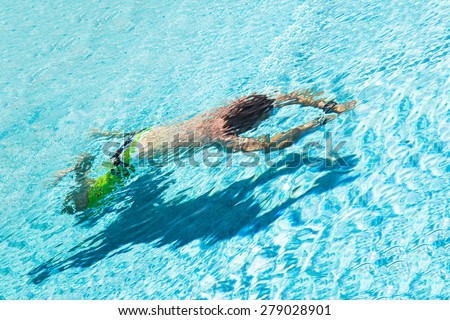 A man wearing green trunks swimming under water. There is a shadow of his body on the bottom of the pool.
