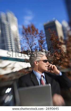 Prime adult Caucasian man in suit sitting at patio table outside with laptop talking on cellphone wearing sunglasses with buildings in background.