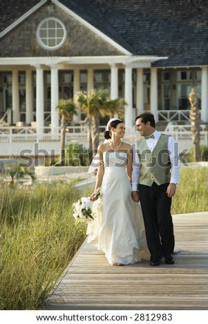 Caucasian mid-adult bride and groom walking down wooden beach walkway holding hands.
