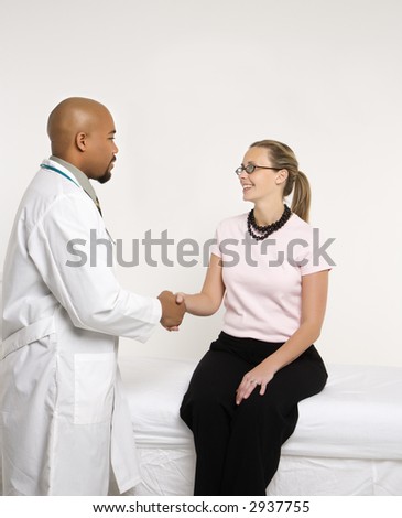 Mid-adult African-American doctor and Caucasion mid-adult female patient shaking hands in doctor\'s office.