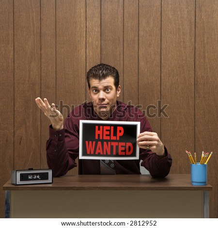 Caucasion mid-adult retro businessman sitting at desk holding help wanted sign with pleading expression.