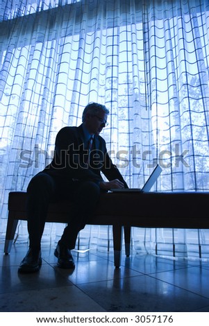 Blue tone silhouetted image of prime adult Caucasian man in suit sitting on bench typing on laptop.