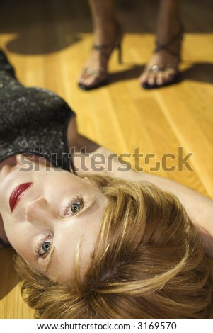 Caucasian mid adult woman lying on wood floor looking at viewer with feet of another woman in background wearing heels.