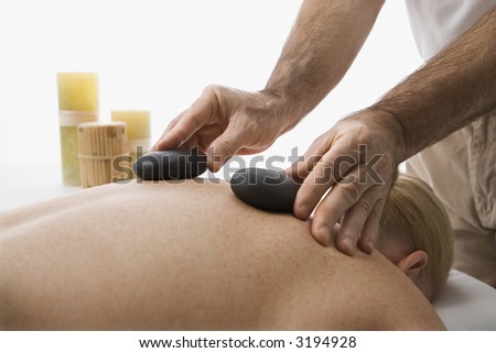 Caucasian middle-aged male massage therapist placing hot stones on back of Caucasian middle-aged woman lying on massage table.