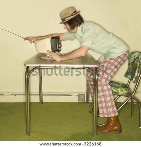 Side view of Caucasian mid-adult man wearing hat and plaid pants leaning over 50\'s retro dinette set adjusting old television set.