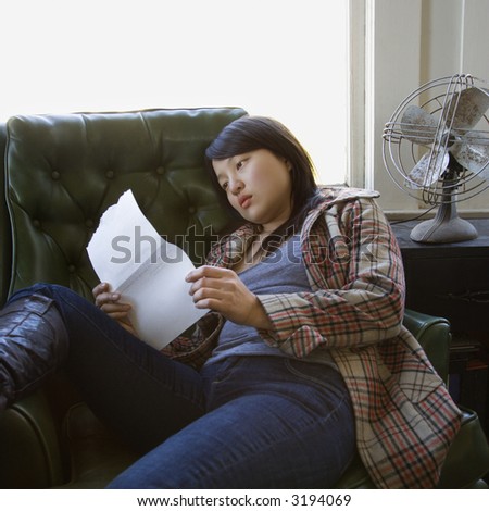 Pretty young Asian woman sitting in green chair reading paper.